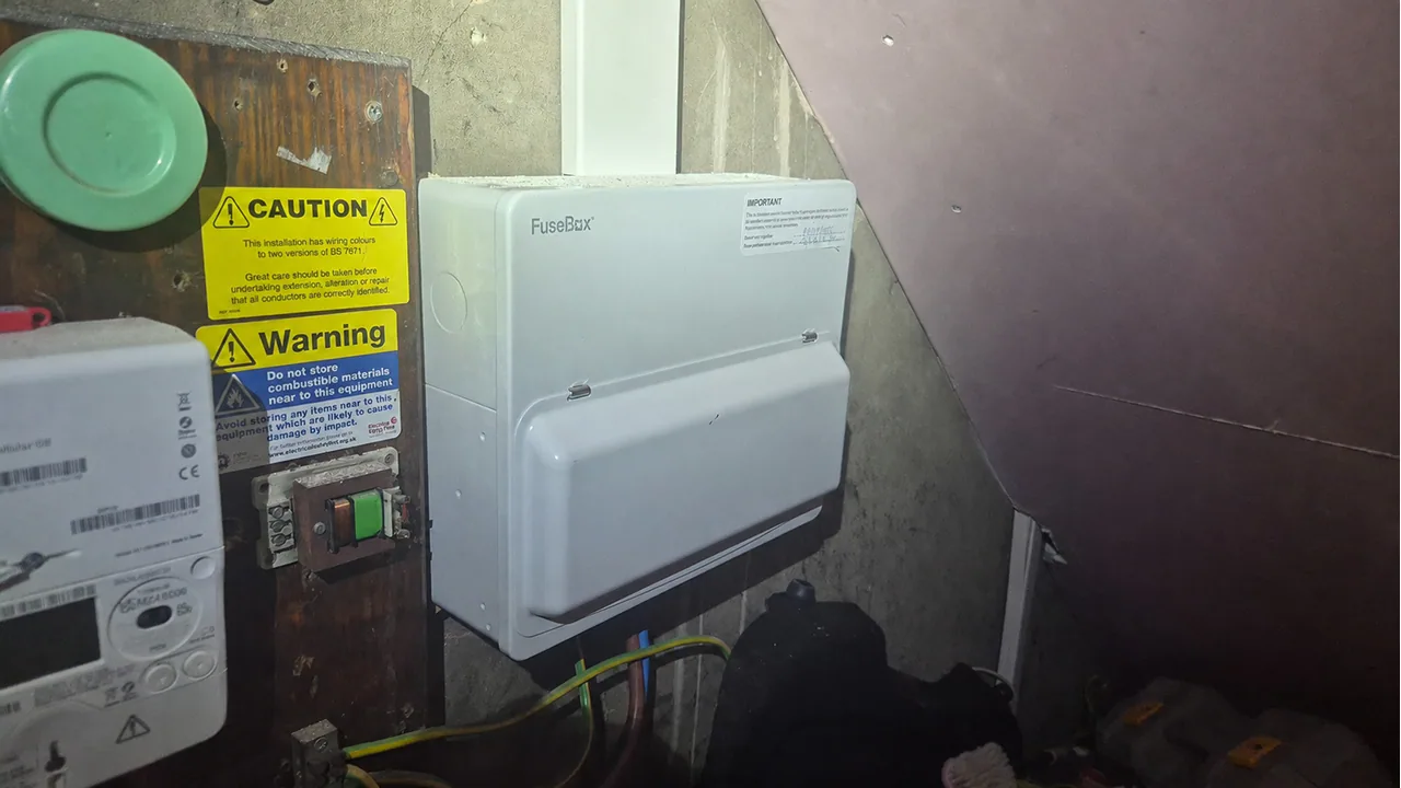 Closed FuseBox consumer unit beside electrical caution labels in an under-stairs cupboard.