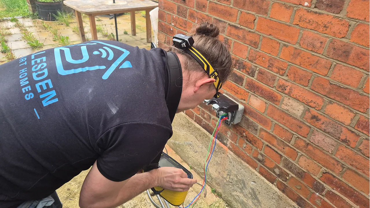 Electrical testing of an outdoor socket circuit with EICR test leads on a brick wall.