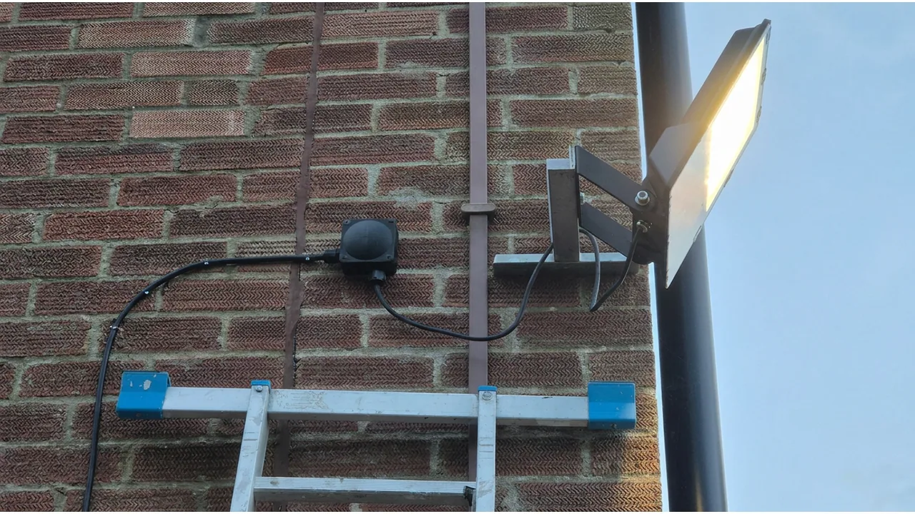 Lit outdoor floodlight with a motion sensor and surface cabling on a brick wall.
